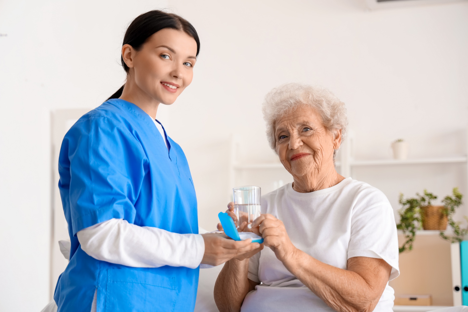 A healthcare professional in a blue scrub top offers a glass of water and a pill to an elderly woman in a white t-shirt. The scene appears to be in a clean, well-lit room, possibly a medical facility or a home setting, with shelves in the background.