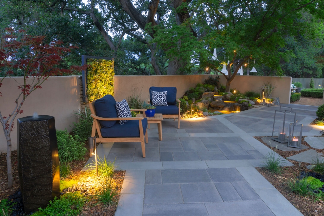 Serene outdoor patio at dusk with blue lounge chairs, stone pavers, warm landscape lighting, lush trees, and a peaceful garden setting.