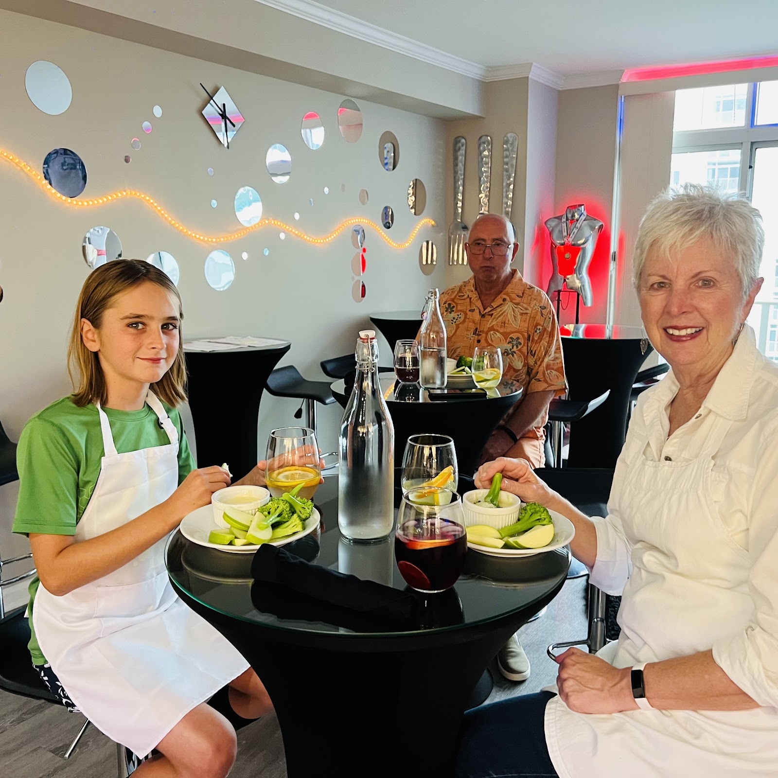 A young girl and two adults are seated at a round black table in a modern, dimly lit room. The girl, on the left, wears a green t-shirt and a white apron and smiles at the camera. She has a plate with broccoli, apple slices, and a small bowl of dip. The woman on the right, also wearing a white apron, is also holding a piece of broccoli to dip. An older man in a tropical-print shirt is seated in the background. The wall behind them is decorated with circular mirrors and a string of orange lights. A decorative sculpture of a torso with a medal is visible on the right side of the room, illuminated by red light. The table is set with glasses of drinks, a water bottle, and napkins. The overall ambiance suggests a casual dining or culinary event.
