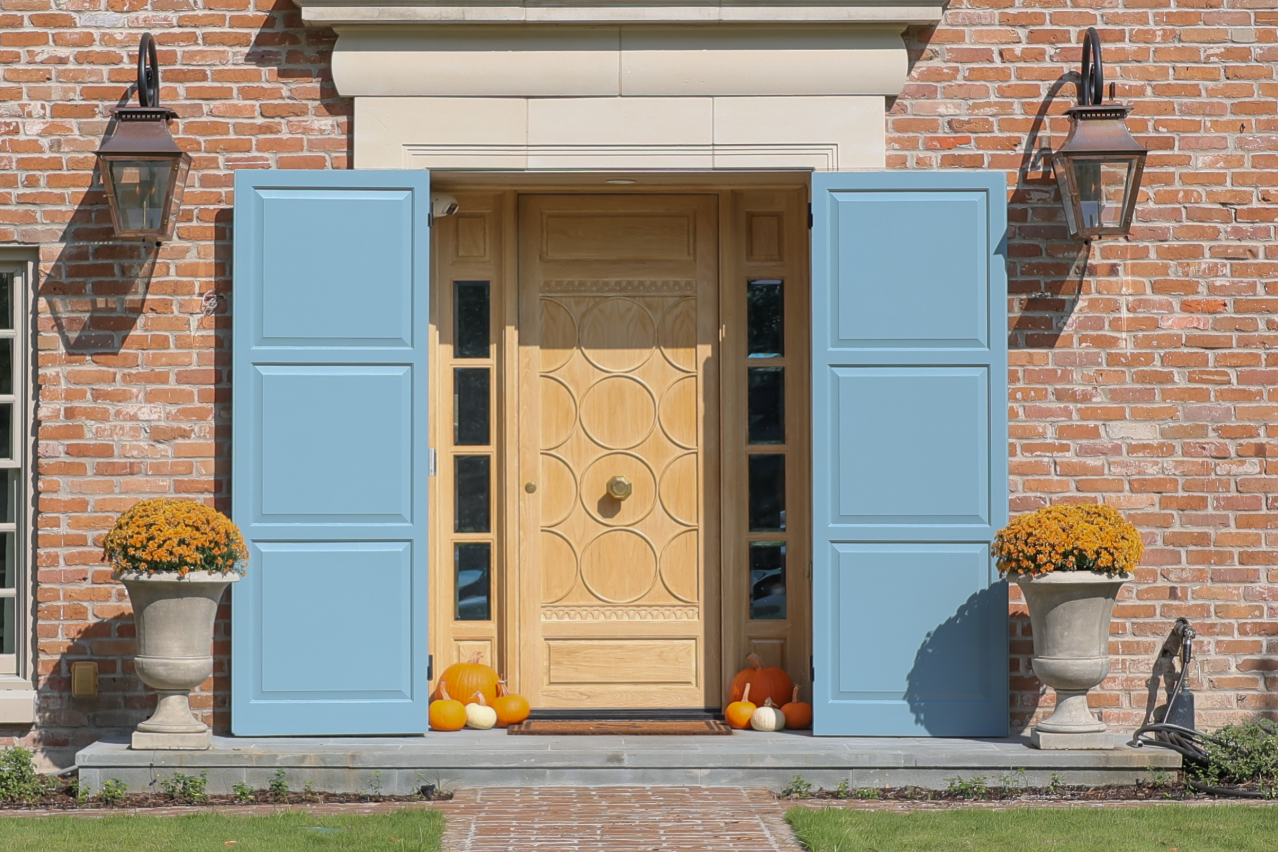 An elegant brick house entrance featuring a light wood door with intricate circular patterns, with two blue doors on each side. Two large urn planters with vibrant orange chrysanthemums stand on either side of the doorway. Small pumpkins in orange and white are arranged at the base of the door, adding a seasonal touch. Traditional wall-mounted lanterns provide additional charm.