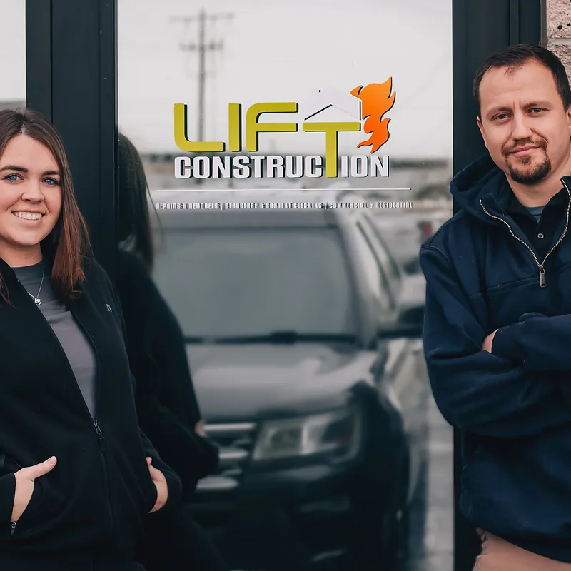 A man and a woman stand in front of a glass door with a construction company logo and text. The woma 