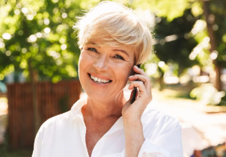 Outdoor portrait of a smiling woman with short blonde hair speaking on a mobile phone with lush green trees in the background.
