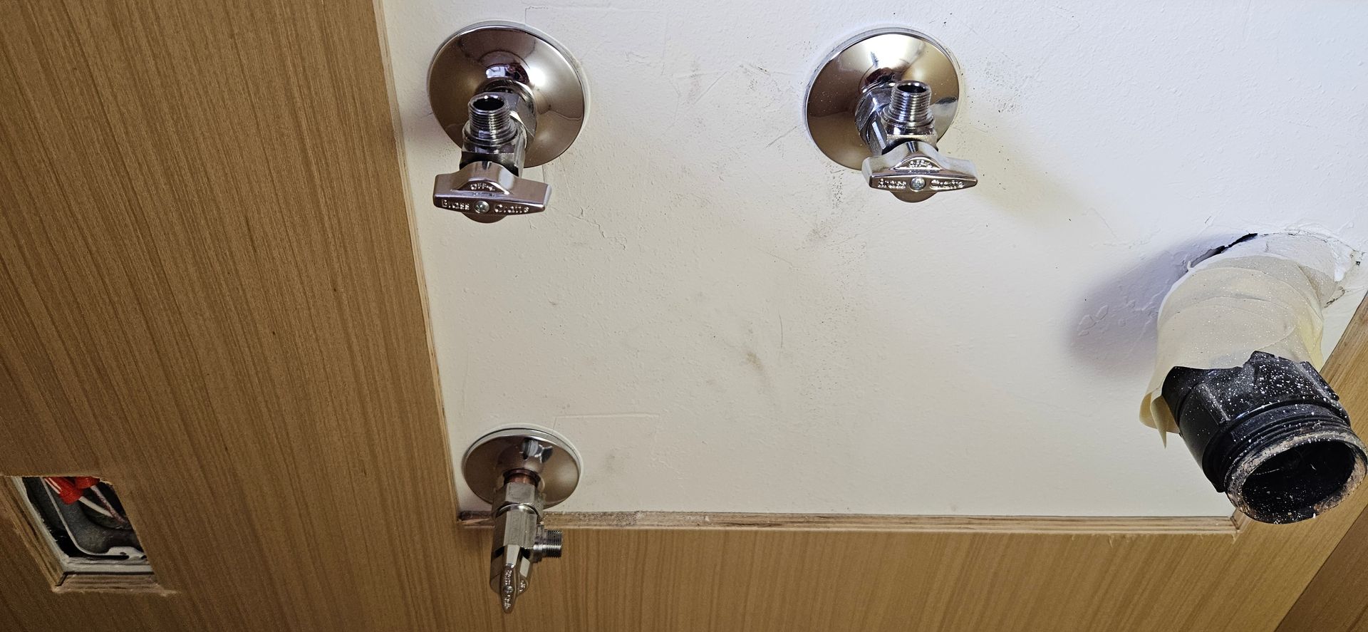 Under-sink plumbing setup showing three chrome shutoff valves mounted on the wall and a black drain  