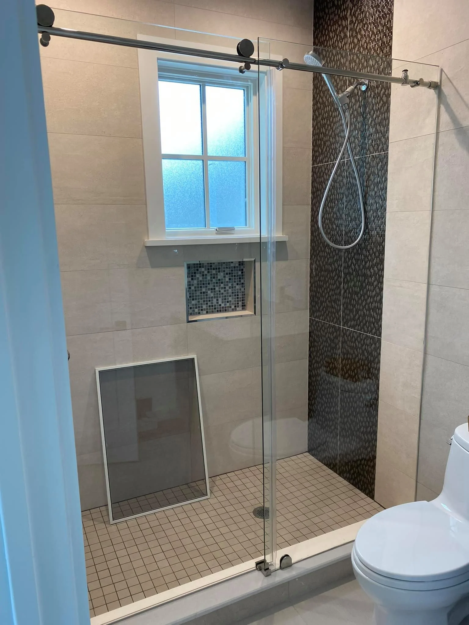 A modern bathroom featuring a sliding glass shower door with a barn door style track, a tiled shower stall with a niche and shower head, a frosted window above the niche, and a white toilet in the foreground. A screen is propped up on the shower floor.