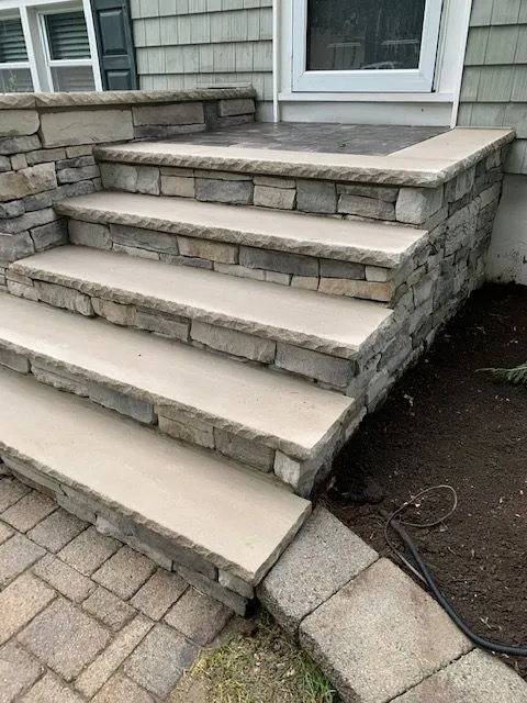 A newly constructed set of stone steps leading up to a house entrance. The steps are made of light-colored sandstone treads with stacked stone risers and a stone retaining wall on the right. The surrounding area includes a paver walkway at the bottom and dark soil with some grass and landscaping in the foreground and on the right.