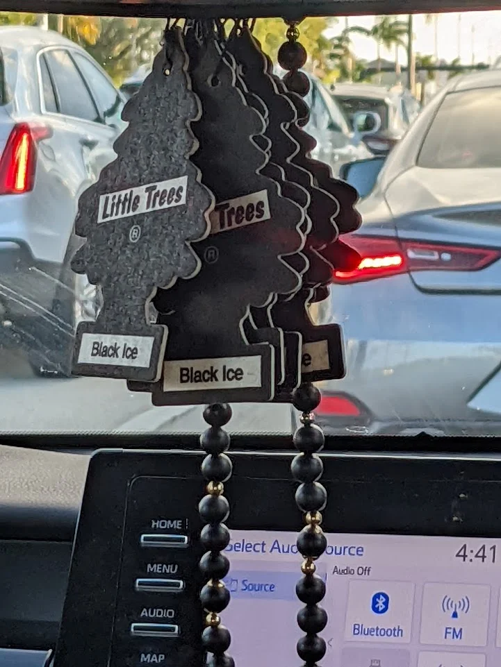 A close-up shot of several black Little Trees air fresheners, specifically the 'Black Ice' scent, hanging in a car. In the background, other vehicles are visible on a sunny day, likely in traffic. The car's dashboard with a media screen is also in the foreground.