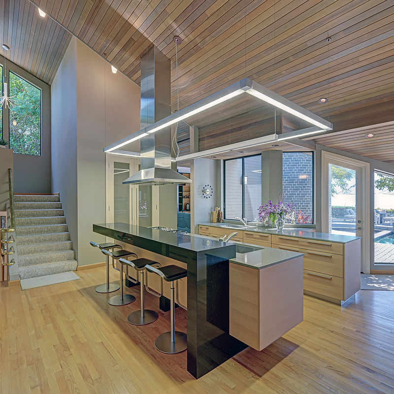 A modern kitchen with a large island, sleek stainless steel appliances, and a wooden ceiling. The island features a black countertop and four bar stools. Large windows offer a view of the outdoors, and a staircase is visible to the left.