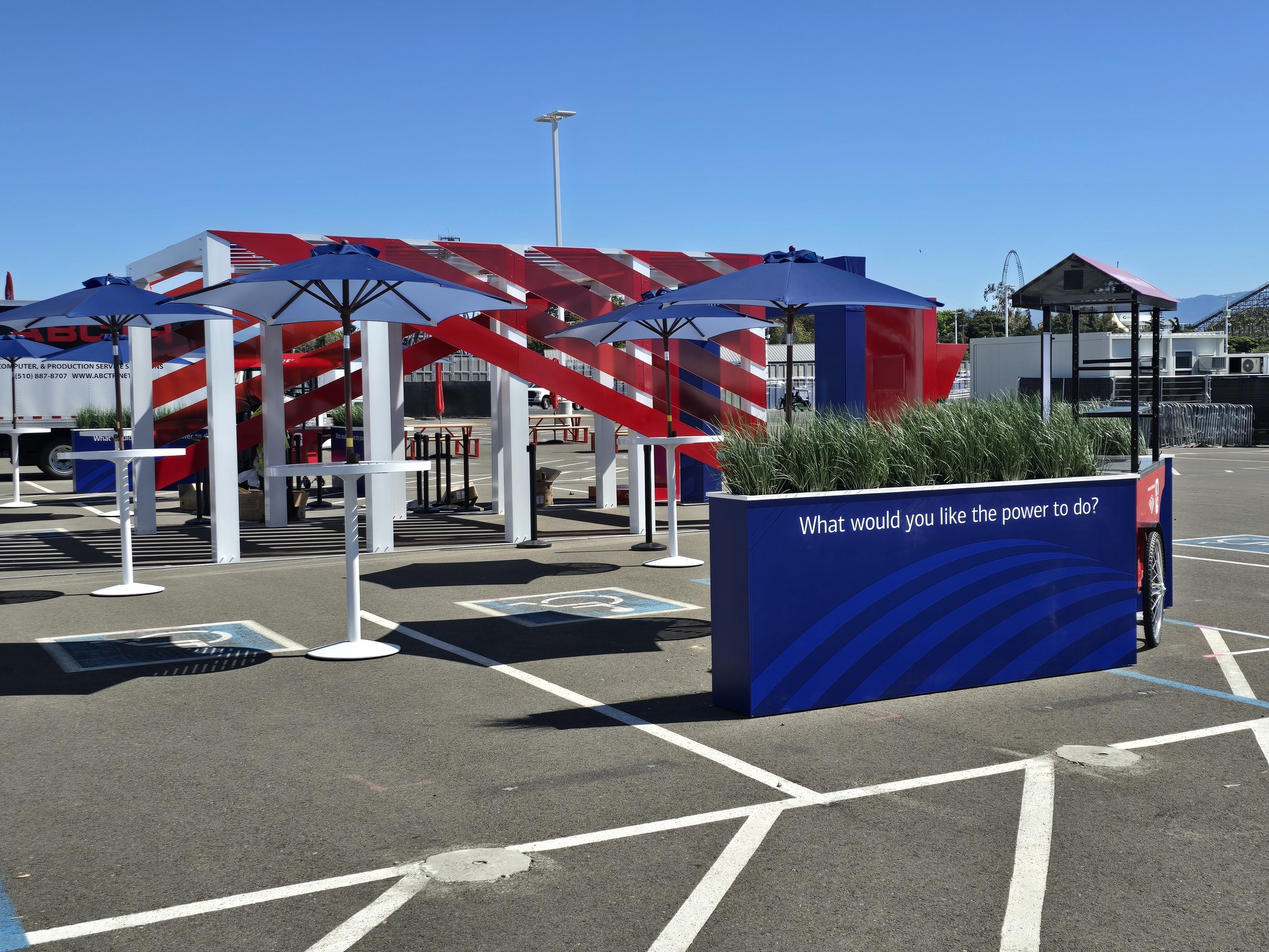 Custom outdoor experiential display featuring a red-and-white architectural structure, branded planter wall, event umbrellas, and modern gathering areas.