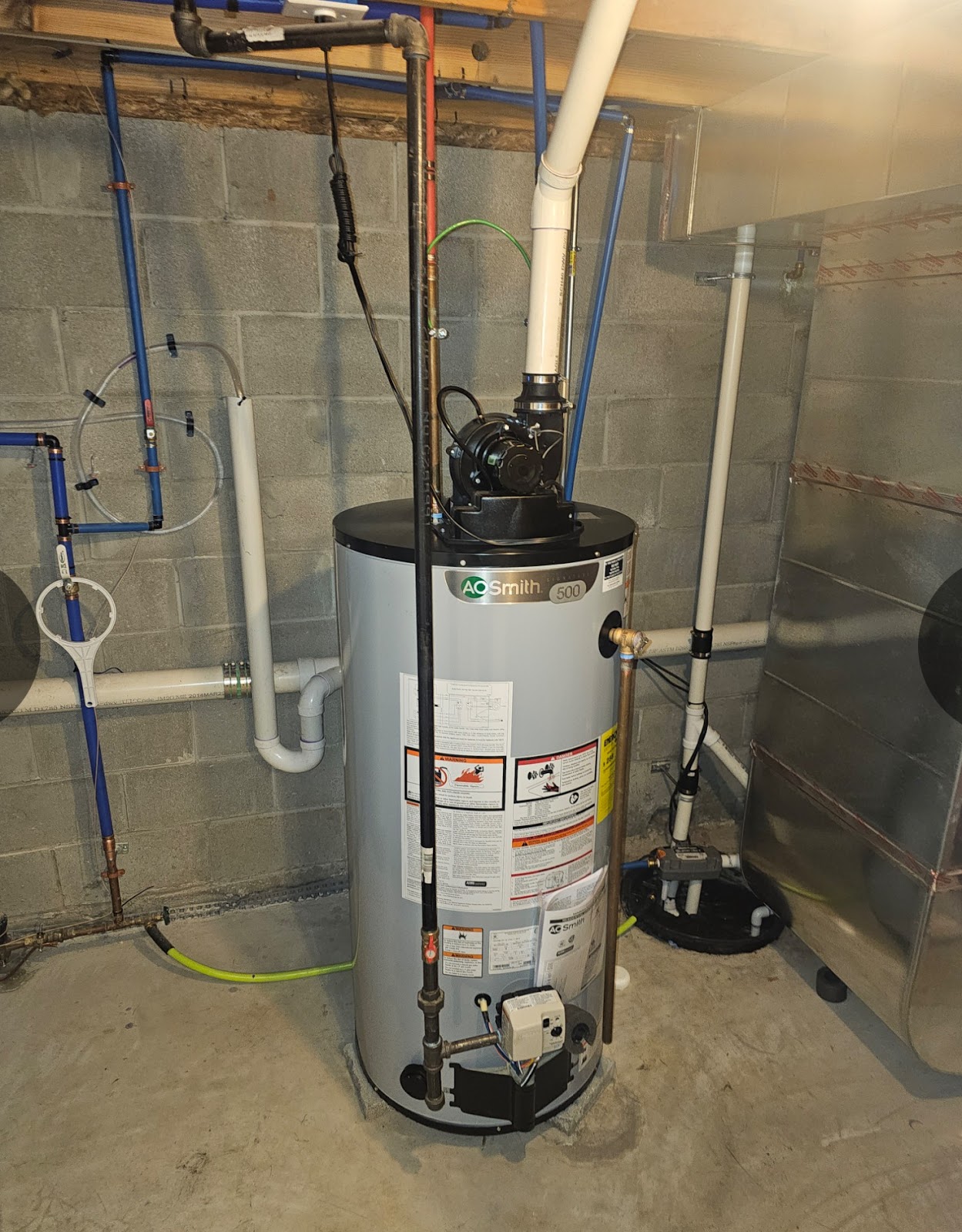 A commercial water heater with an AO Smith logo and model number '500' is prominently displayed in a basement or utility room. The water heater is surrounded by various pipes, including blue PEX tubing, black gas lines, and white PVC exhaust pipes, all connected to the unit. To the right of the water heater, a large metal ductwork is visible, and a sump pump is located at the base of the water heater. The walls are made of concrete blocks, and the floor is concrete.