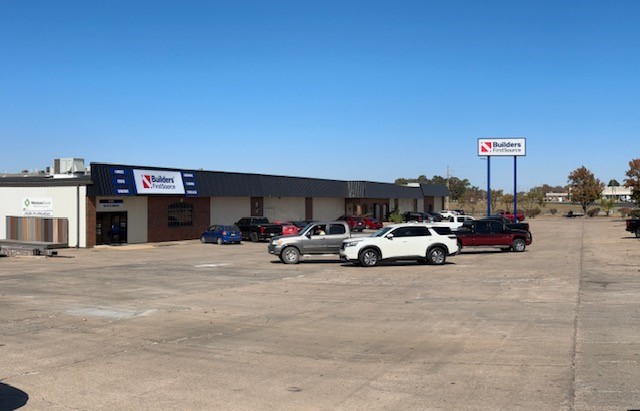 A wide shot of a Builders FirstSource facility with a parking lot in the foreground. The building has a mix of brick and metal siding with large signage displaying the Builders FirstSource logo above the entrance. Several cars and trucks are parked in the lot. To the right, a large standalone sign with the Builders FirstSource logo is visible near the street. The surrounding area includes trees and other commercial buildings under a clear blue sky, indicating a sunny day.