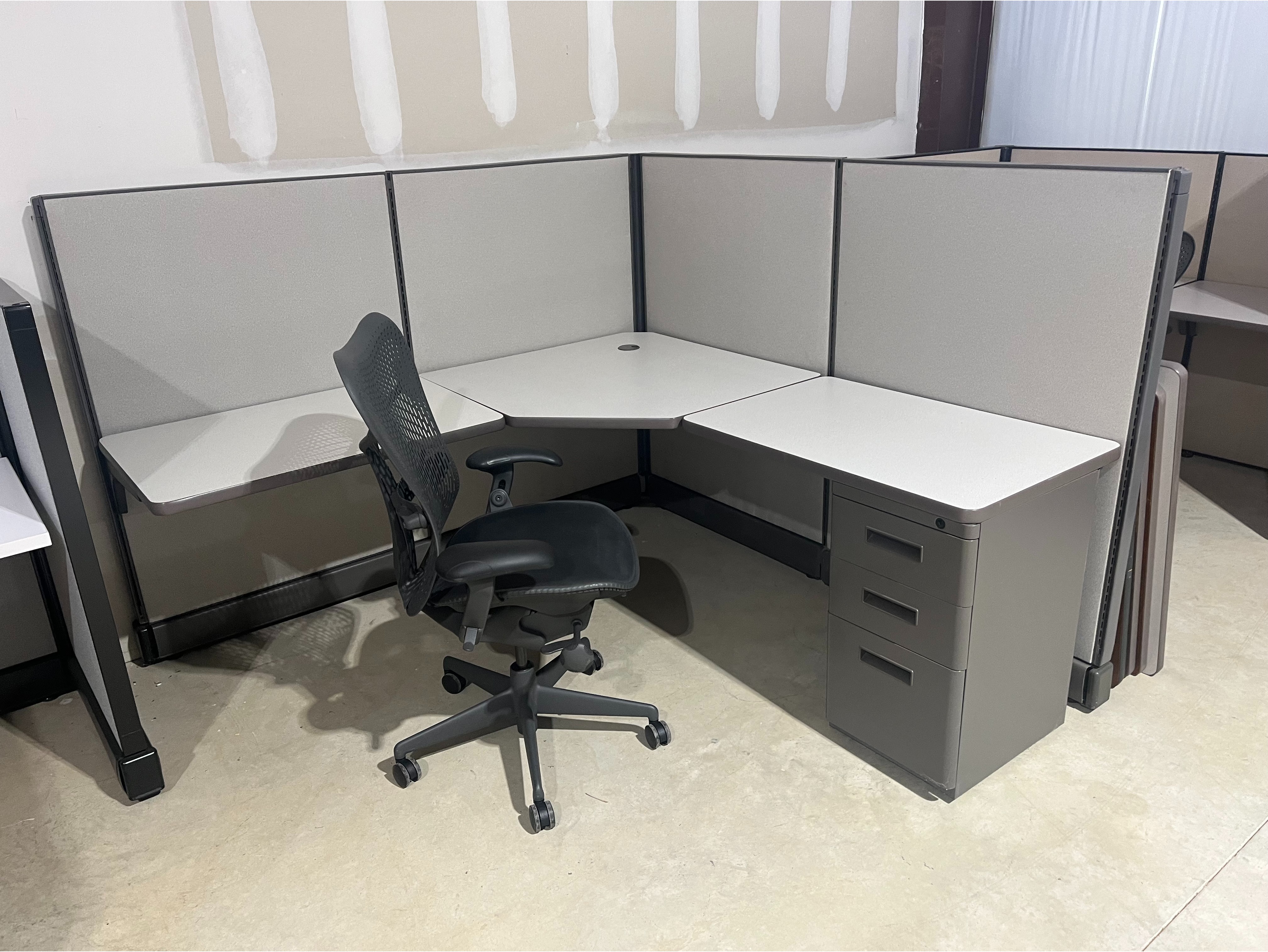 Modular Corner Workstation:

Modern L-shaped office cubicle featuring gray fabric privacy panels, an ergonomic black office chair, and a spacious light-oak desk surface.