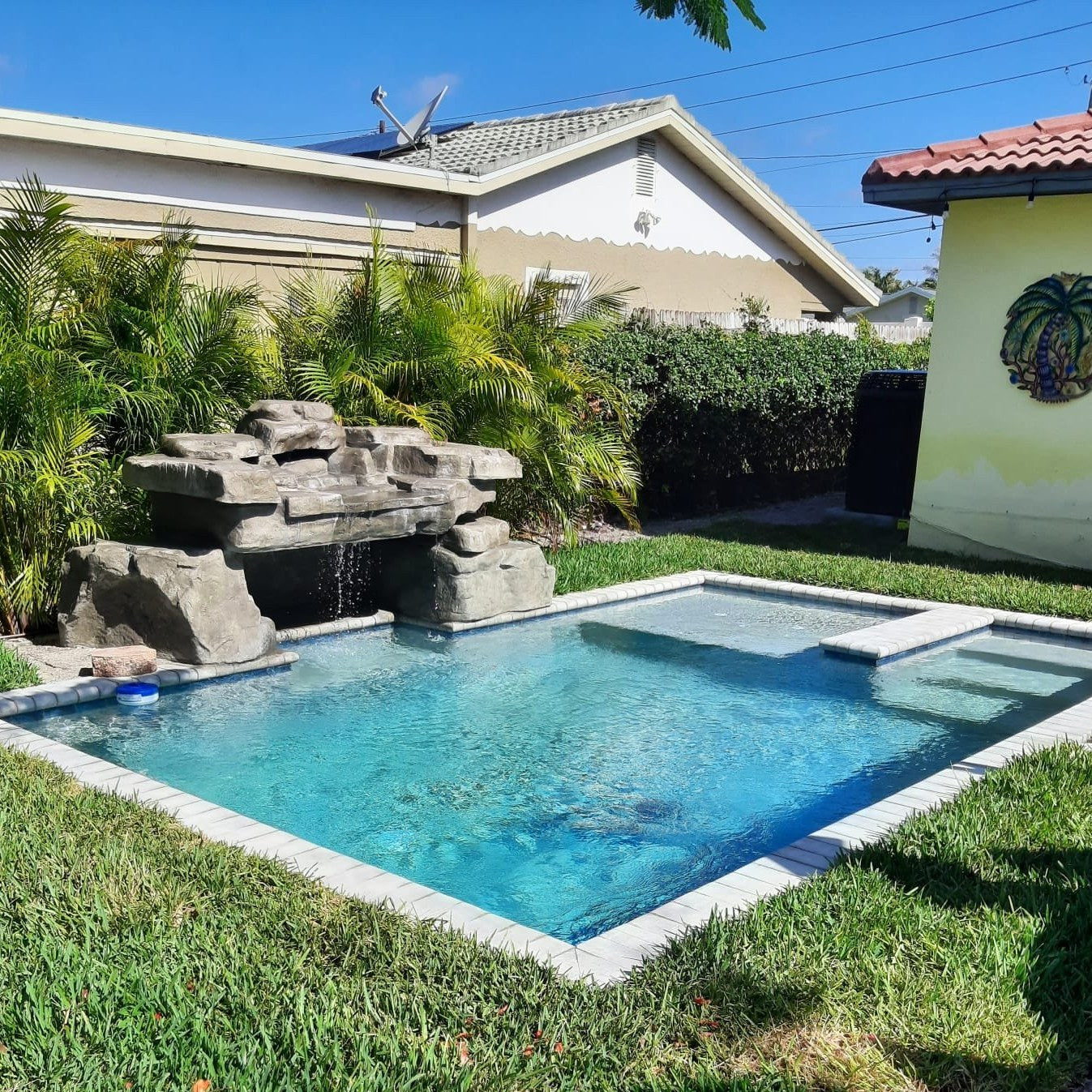 Residential backyard pool and spa with a natural rock waterfall feature, surrounded by tropical landscaping and green lawn.