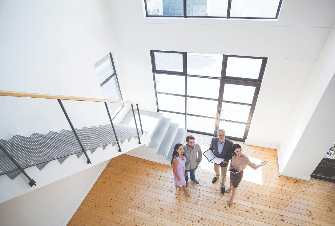 Aerial view of three people with a real estate agent inside a bright, spacious room with a wooden floor and large windows, suggesting a property tour.