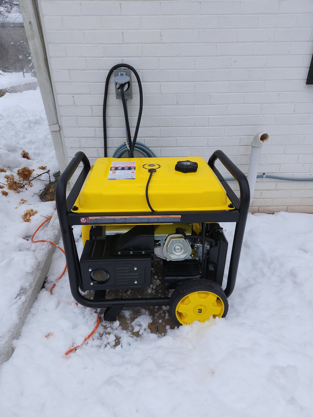 A bright yellow generator sits in the snow outside a brick building, plugged into an outdoor outlet.