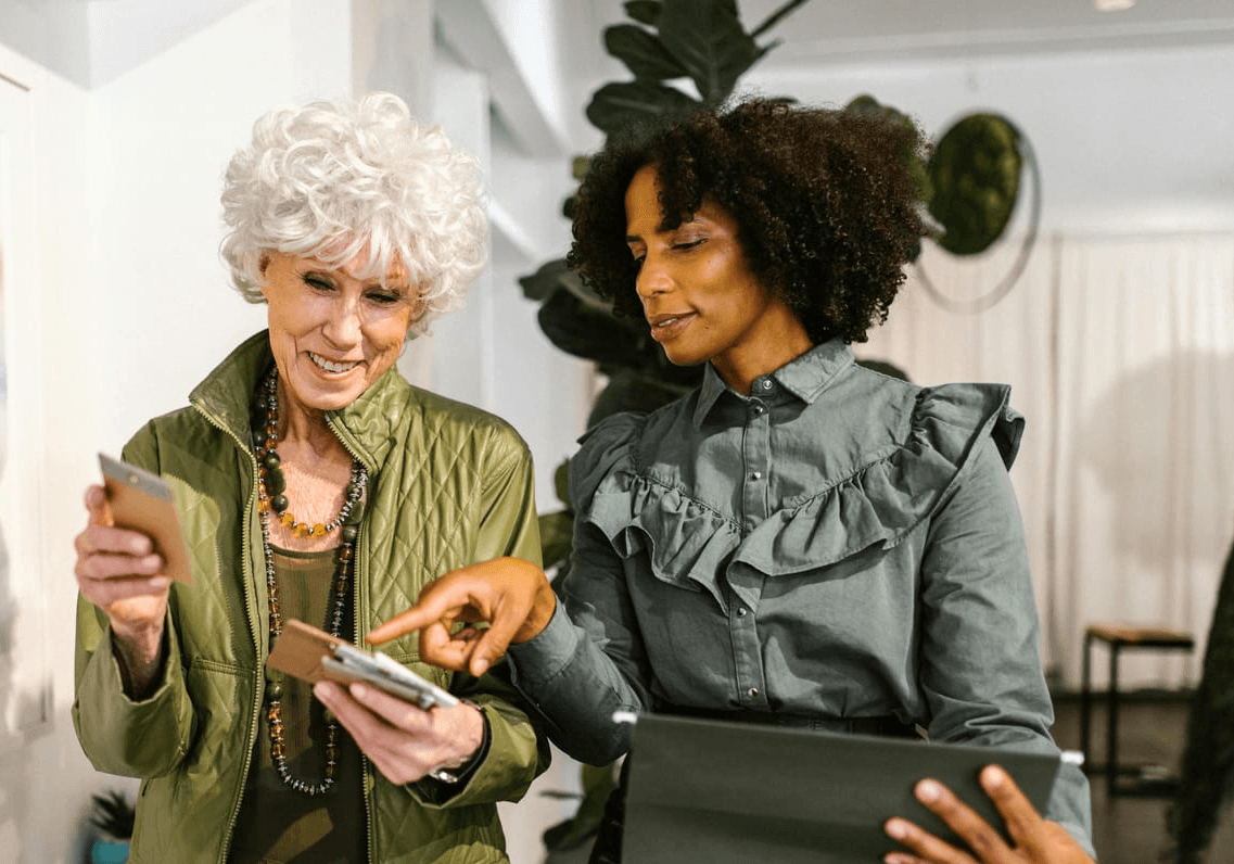 Two women, one older with white curly hair and the other younger with dark curly hair, are looking at a tablet device. The older woman is wearing a green quilted jacket and a necklace. The younger woman is wearing a grey button-down shirt with ruffles on the chest and shoulders. They appear to be in a store or office setting with plants and curtains visible in the background.