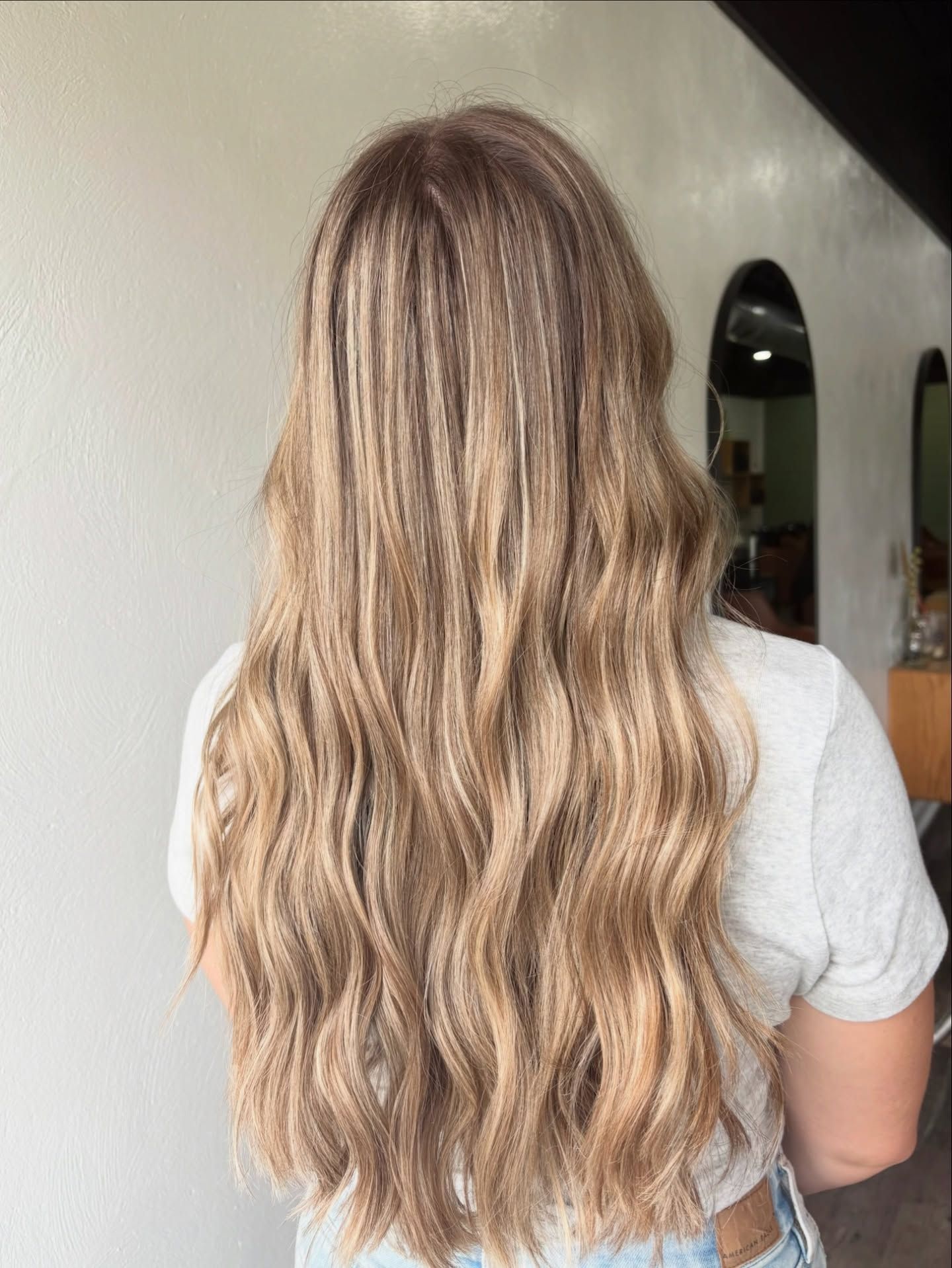Back view of woman with long wavy blonde hair featuring natural highlights, styled softly inside a modern hair salon.