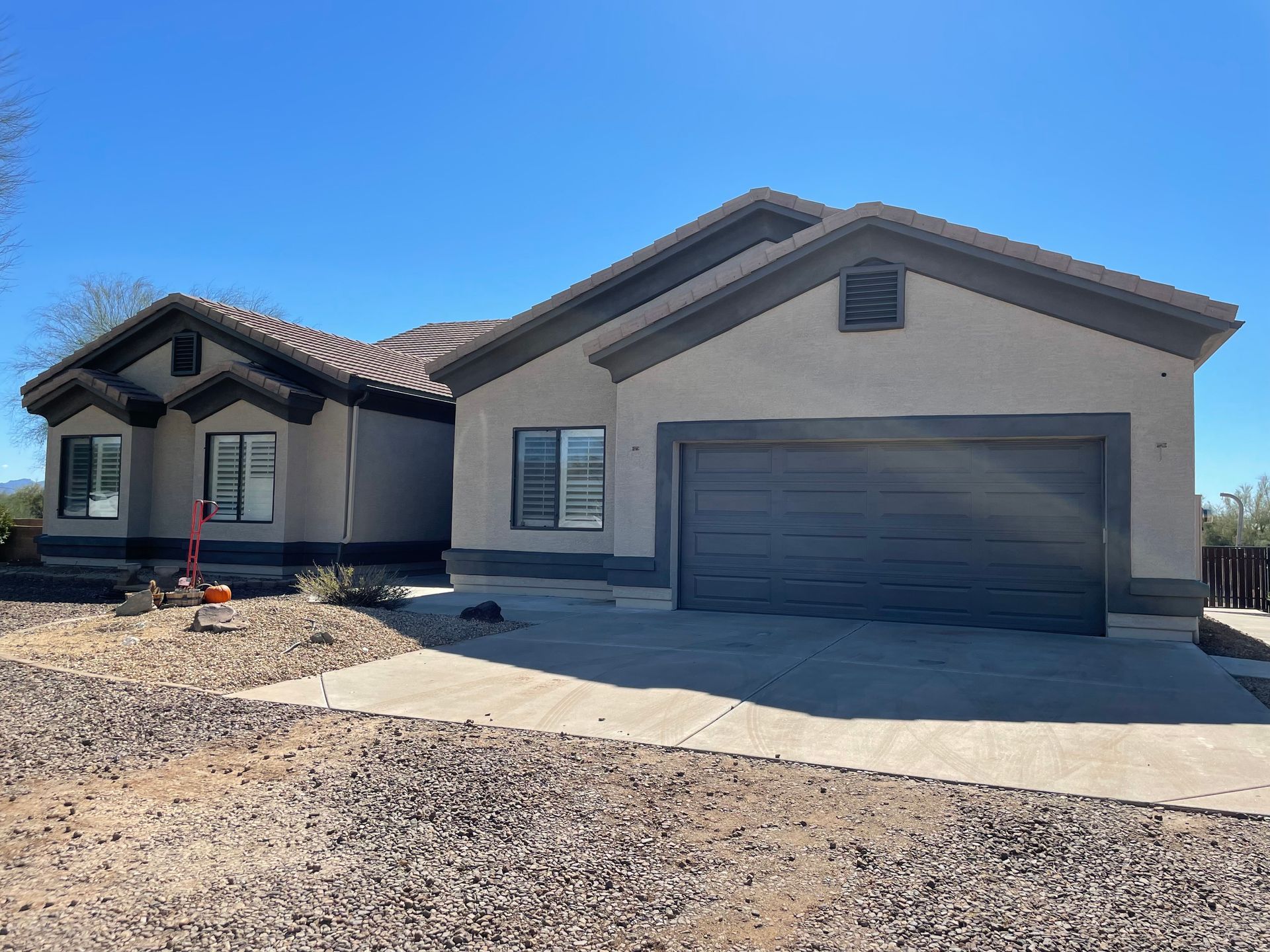A modern single-story house with a two-car garage, stucco exterior, and desert landscaping sits under a clear blue sky, featuring clean architectural lines and a spacious driveway.