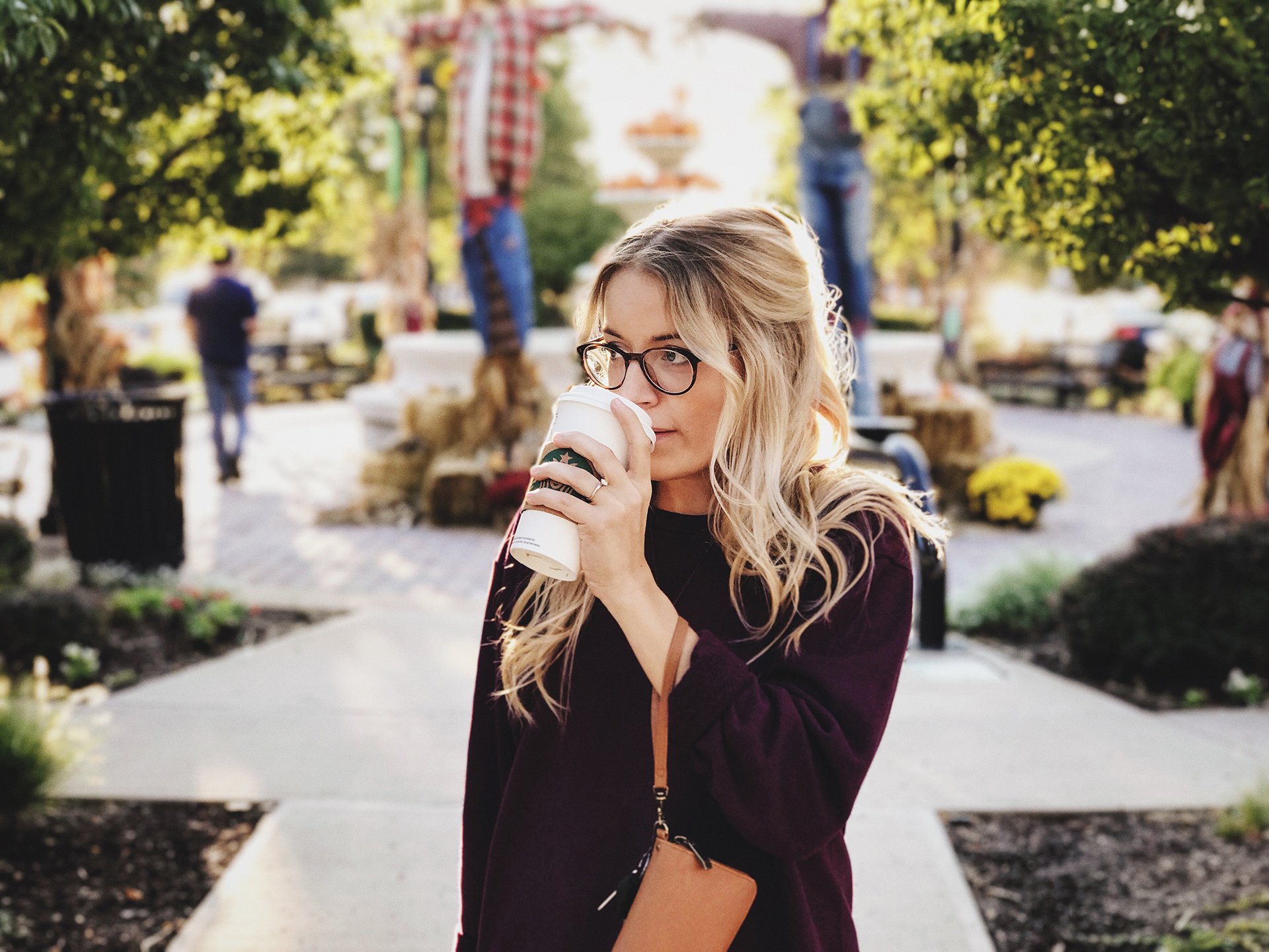 A blonde woman wearing glasses and a burgundy sweater drinks from a Starbucks coffee cup. She is outdoors with scarecrows and trees in the background, suggesting an autumn or harvest festival setting.