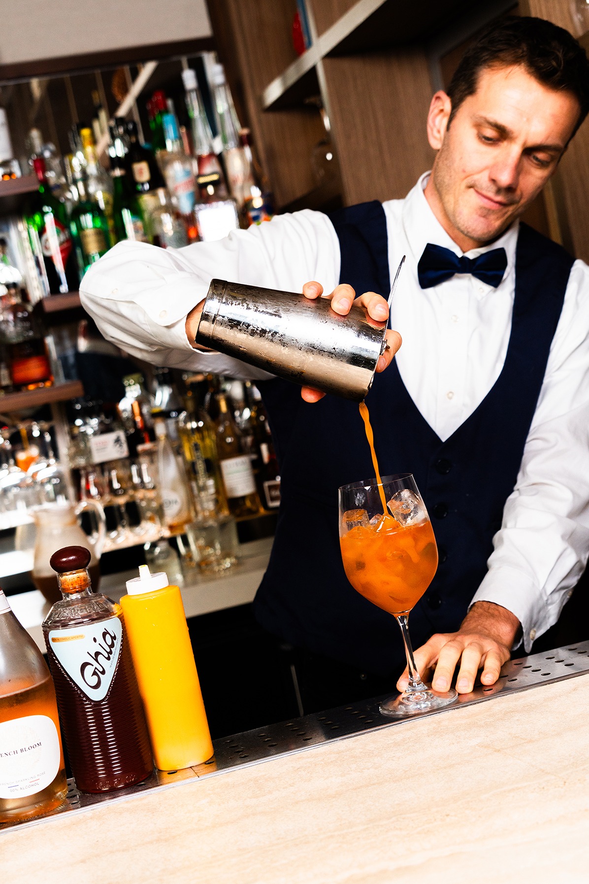A skilled mixologist expertly prepares an Italian spritz at a well-stocked bar. Bottles of Ghia and other premium spirits are displayed on the counter in the foreground.