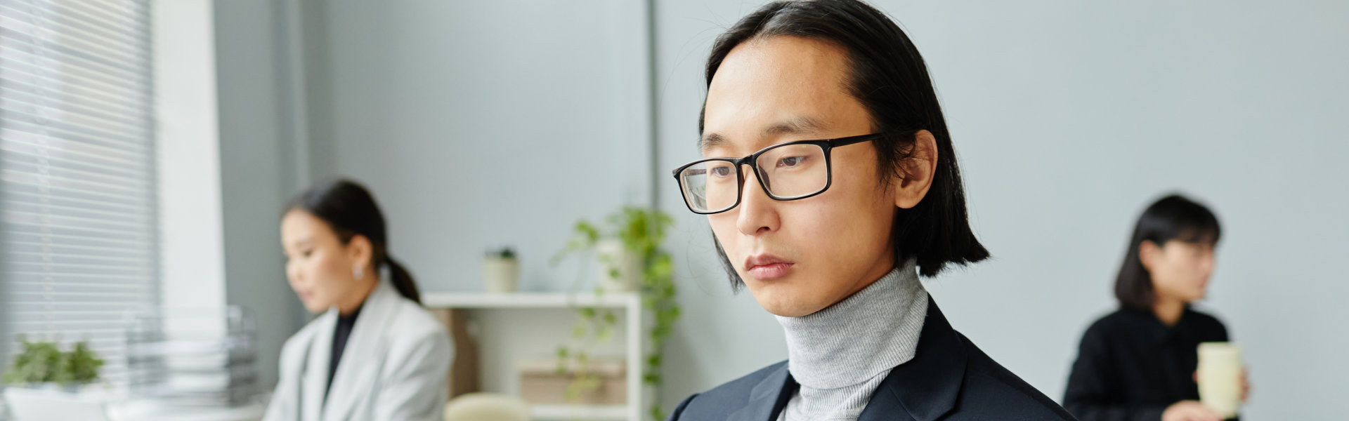 A close-up shot of a man with glasses in an office setting, with two women blurred in the background 