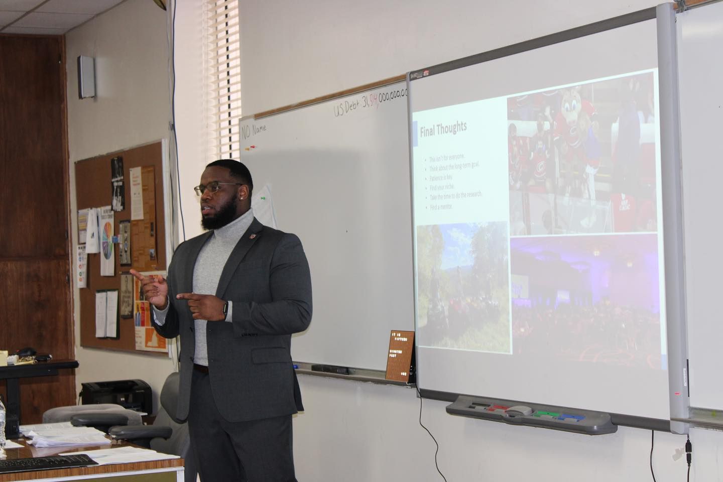 A professionally dressed man gives a presentation in a classroom, gesturing while speaking, with a slideshow projected onto a whiteboard.