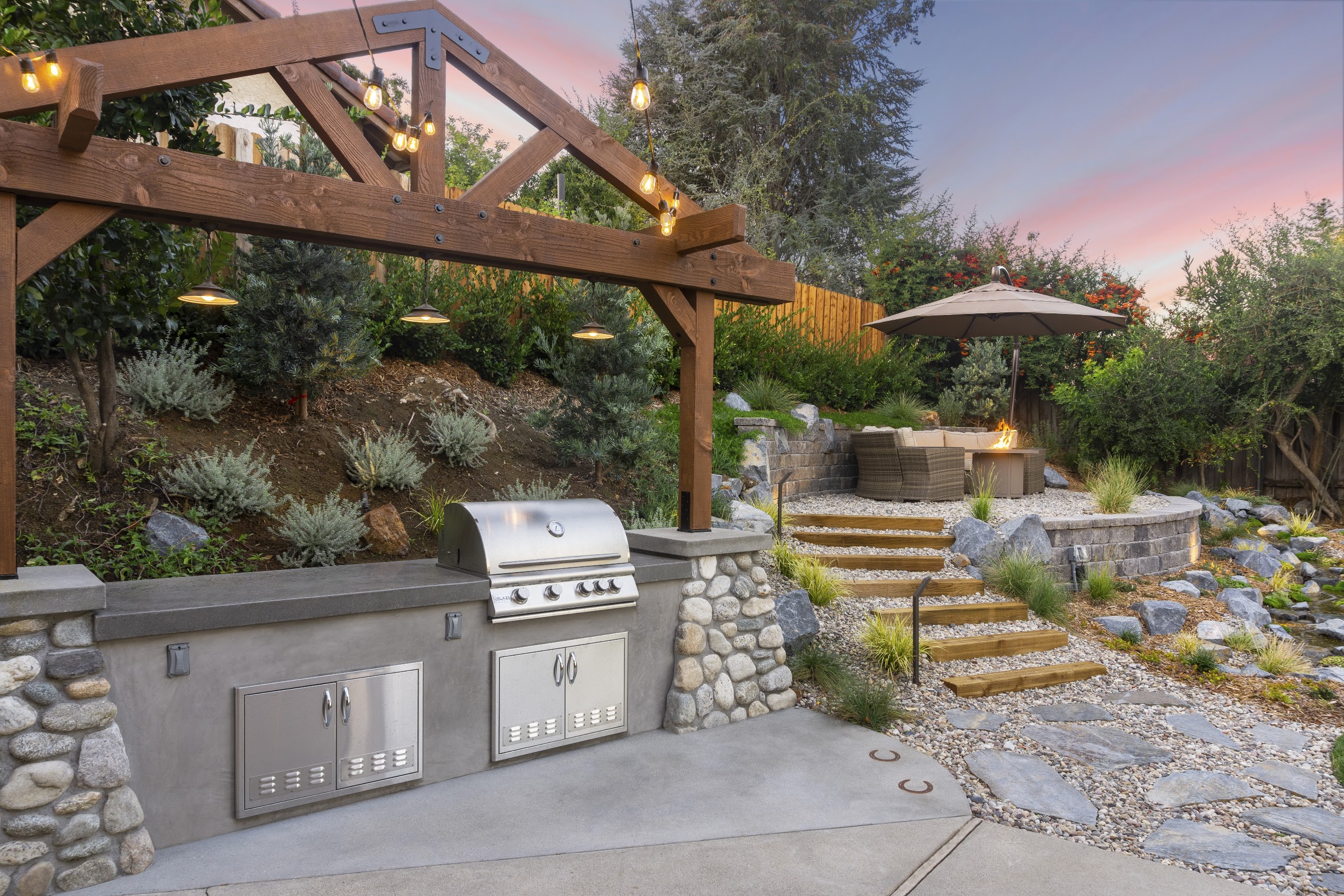 Outdoor backyard kitchen with stainless-steel grill, timber pergola and string lights, stone steps, fire pit, and lush landscaping at sunset.