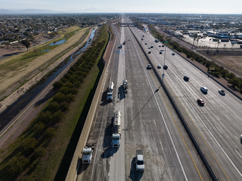 High-altitude aerial drone photograph looking down along a multi-lane divided highway with open desert landscape on the left and commercial development on the right. In the center closed lanes, a full DSI grinding train—including support tankers and grinding machinery—works sequentially along the concrete roadway while active traffic flows in the far right lanes.