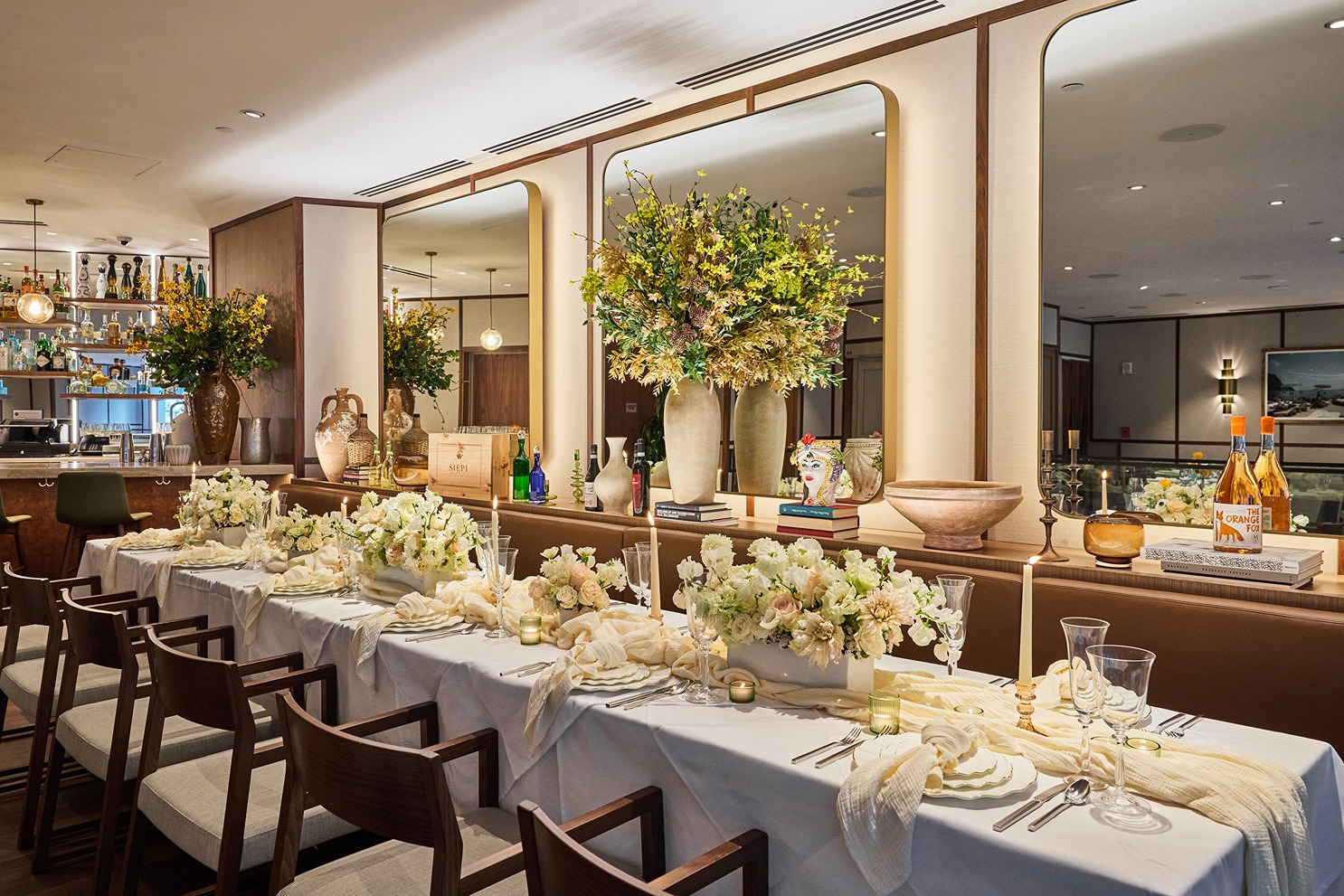 An elegantly decorated long table featuring white linens, floral arrangements, and candles, set against a backdrop of large mirrors and warm lighting.