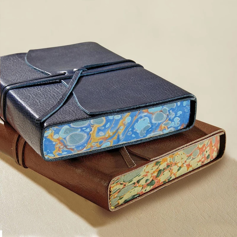 A refined product image by Tabula Rasa Social Stationers, showcasing two Cavallini Journals. The top journal, in navy blue, is layered over a classic brown one. Both embody a timeless charm with their sophisticated leather-like covers and secure flap closures. A highlight of their vintage-inspired artistry is evident in the vibrant, unique marbled patterns on their page edges. These journals combine high-quality craftsmanship with smooth, durable pages, ideal for sketching, notes, or planning, reflecting an appreciation for nostalgic beauty and the joy of writing.