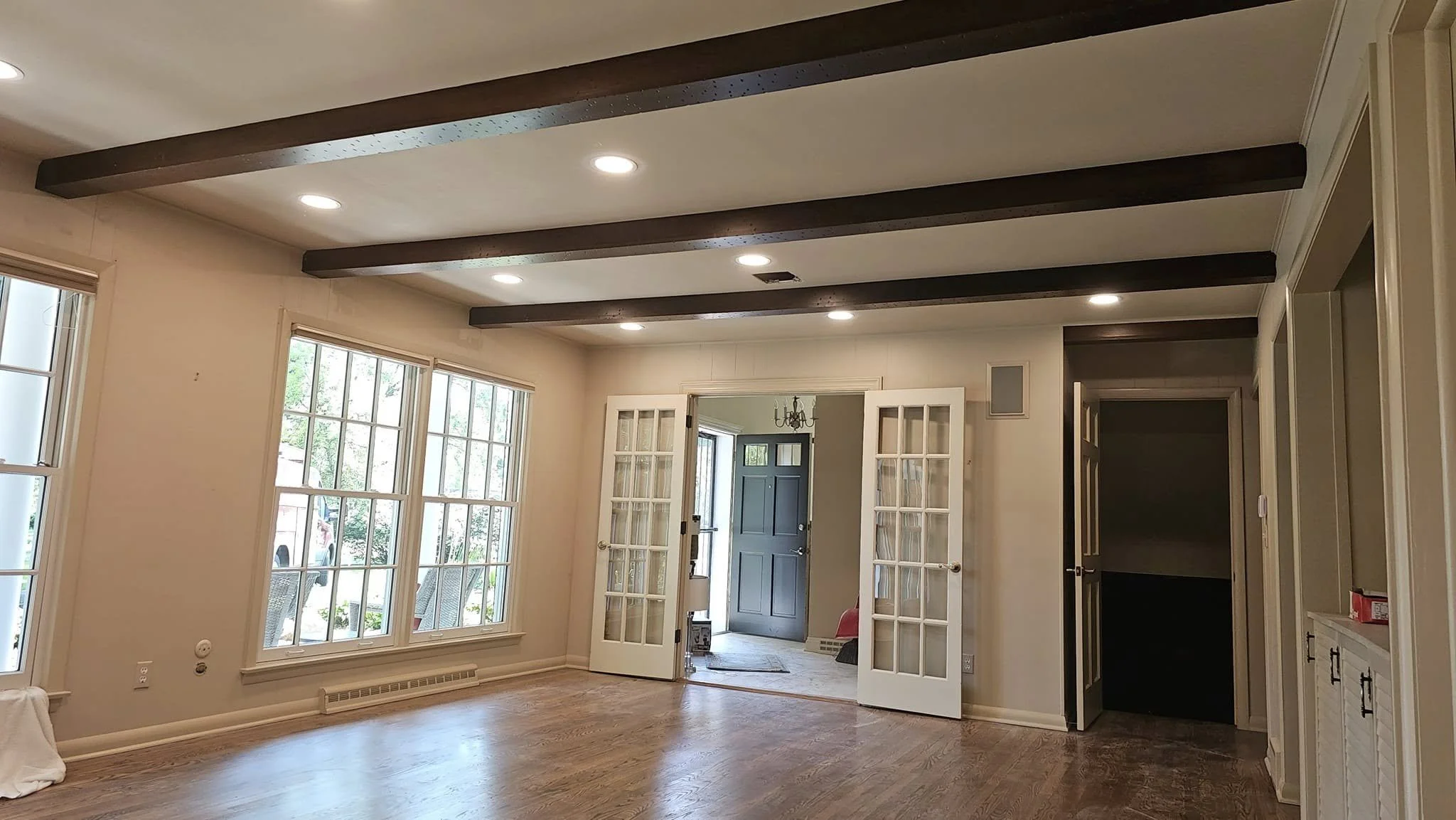 A view of a living room with dark wooden beams on the ceiling, recessed lighting, and large windows. French doors open into another room with a dark blue door at the end of a hallway.