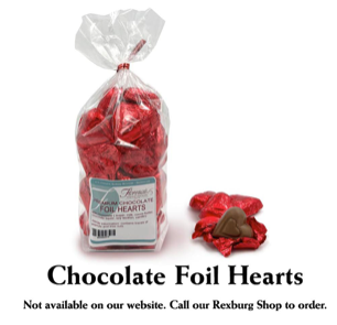 This photograph showcases Florence's Exquisite Chocolates' "Premium Chocolate Foil Hearts," likely from their Rexburg, Idaho location. The primary focus is a clear bag overflowing with red foil-wrapped chocolate hearts, cinched with a festive red bow. The bag's label clearly identifies the product and includes a barcode. Several loose chocolate hearts, still encased in their shiny red foil, are scattered beside the bag, with one partially unwrapped to reveal the smooth milk chocolate inside.