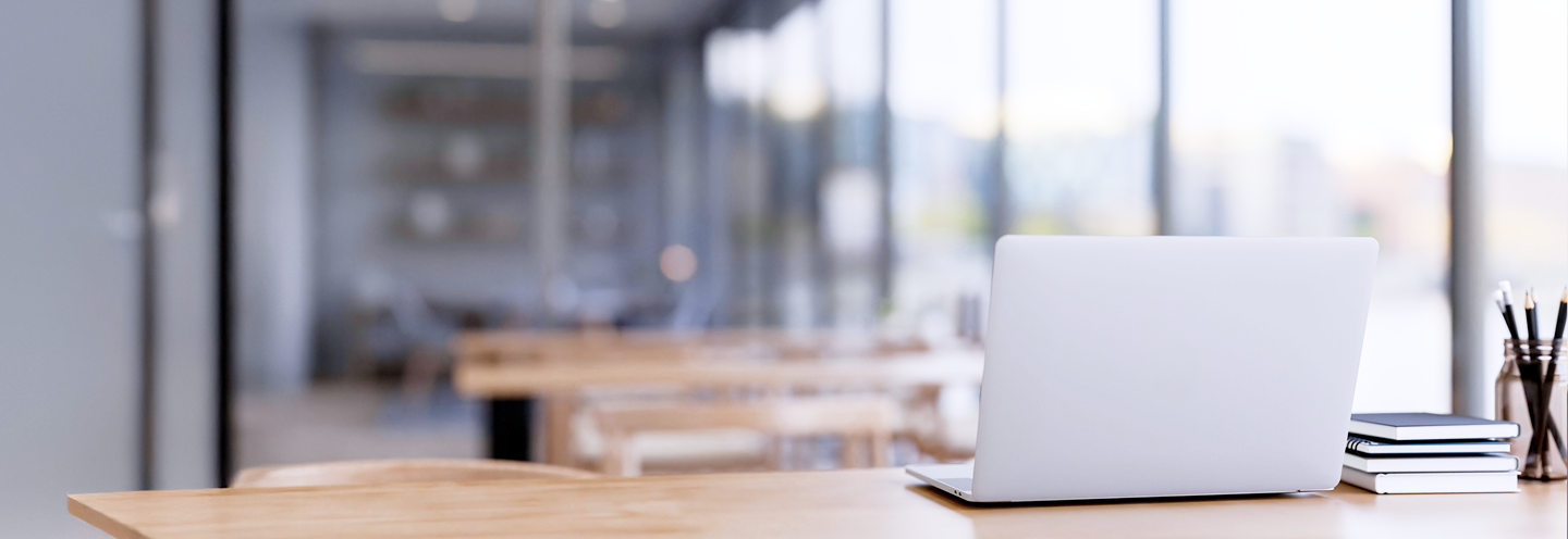 A white laptop on a desk in a study hall with lots of windows.