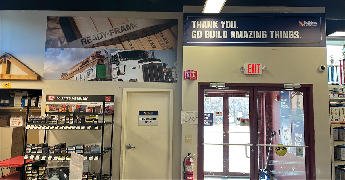 Interior store entrance area featuring a Ready-Frame promotional banner, a Grip-Rite collated fasteners display, and a "Thank You. Go Build Amazing Things." sign above red exit doors leading to the parking lot.