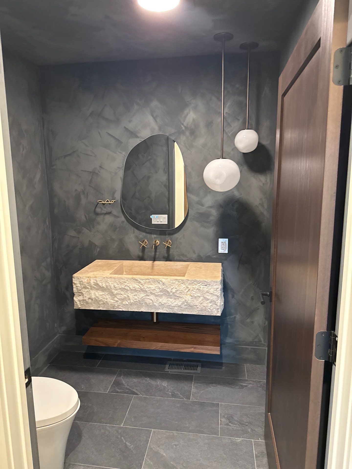 Compact modern powder room features dark textured walls, sculpted stone sink, brass wall-mounted faucet, asymmetrical mirror, hanging globe lights, floating wood shelf, gray tile flooring, and partially visible toilet nearby.