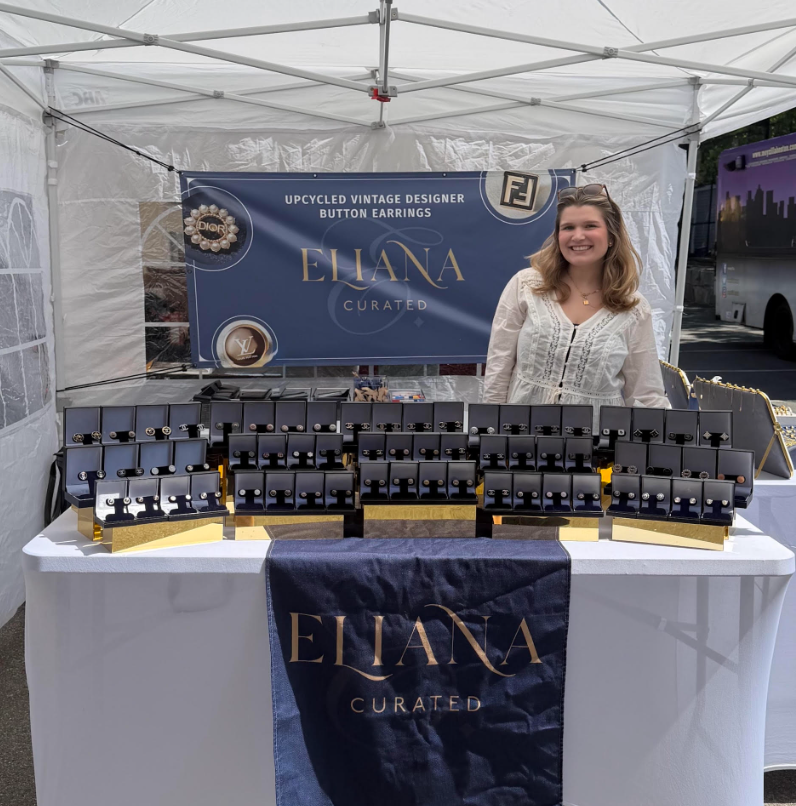 Eliana Curated displays upcycled vintage designer button earrings at an outdoor market booth. Rows of boxed earrings and navy banners highlight unique accessories that give vintage buttons a new life.
