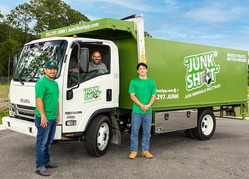 Two Junk Shot team members standing by the front tire of the white Isuzu truck while the driver smil 