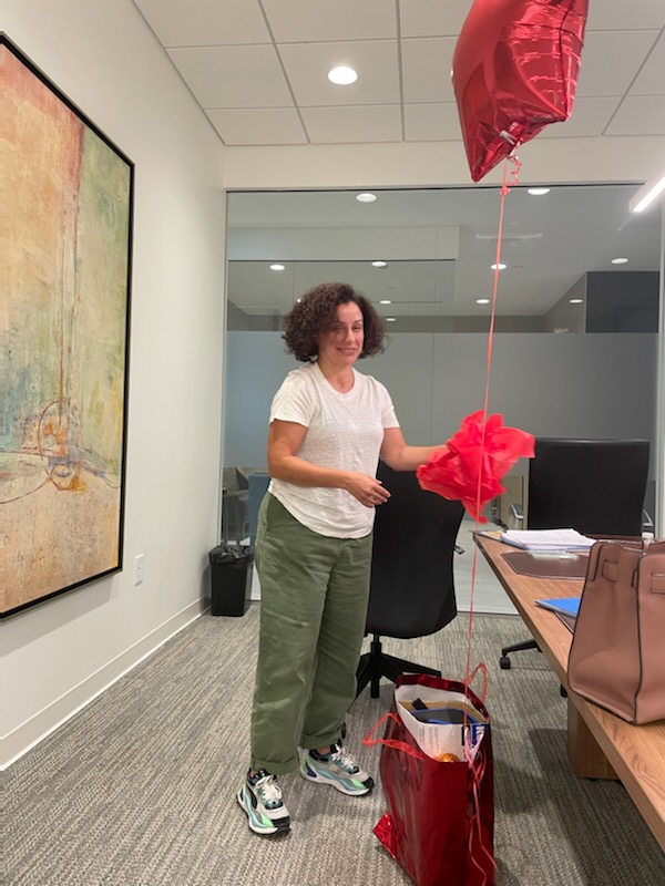A woman in a white t-shirt and green pants stands in an office holding a red balloon. She is holding red tissue paper. A red gift bag is on the floor in front of her, and a pink handbag is on a desk behind her. A painting hangs on the wall to her left.