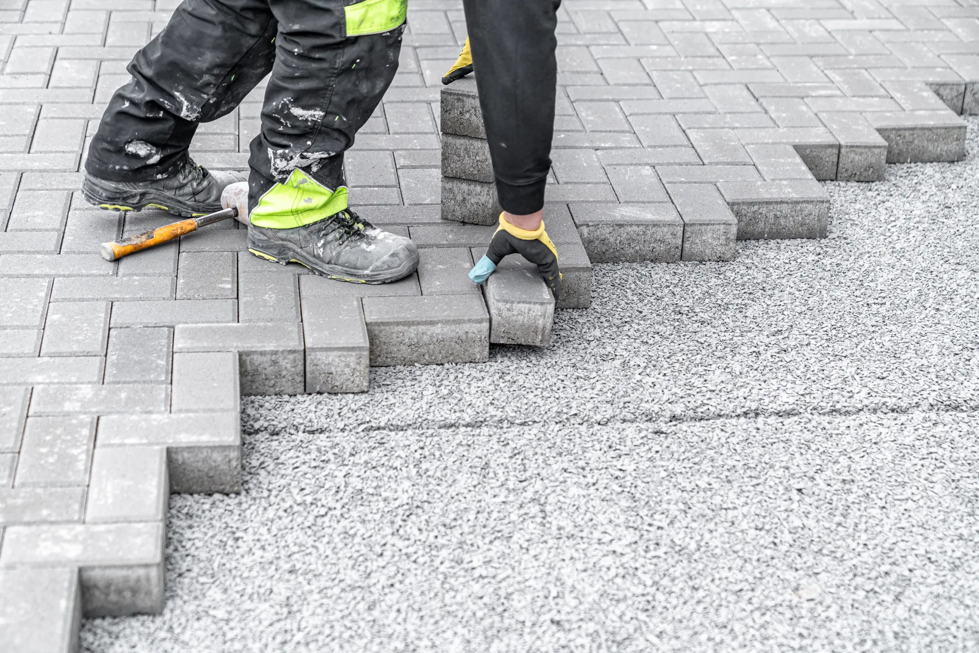 A construction worker, wearing black pants with lime green accents and work boots, is laying down grey paving stones on a bed of gravel. They are using yellow-gloved hands to place a stone and a mallet is visible near their feet.