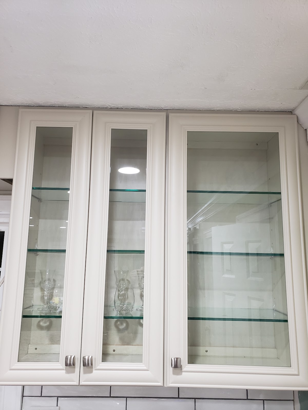A close-up, slightly angled shot of three white kitchen cabinets with glass doors. The cabinets are mounted above a white tiled backsplash. Inside the cabinets, glass shelves hold a few crystal glasses and vases. The ceiling is visible at the top of the image, showing some texture and a light fixture.