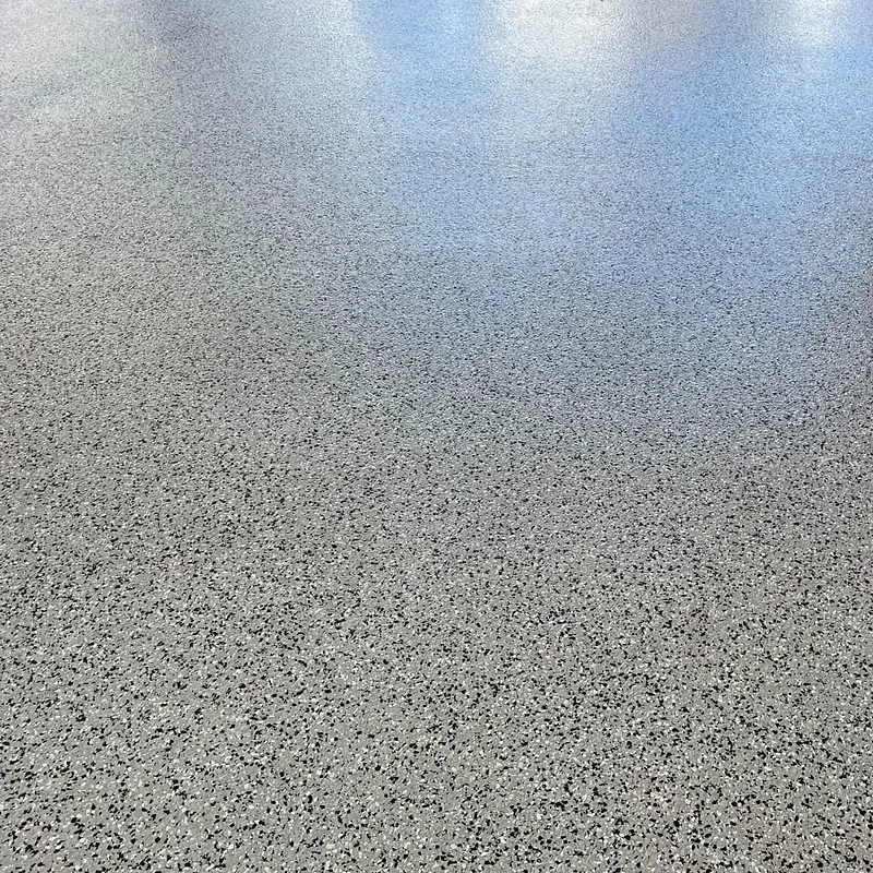 A high-angle close-up shot of a gray speckled floor reflecting the blue sky and white clouds above. The floor appears to be made of a material with small, irregular chips of black, white, and gray embedded in it.