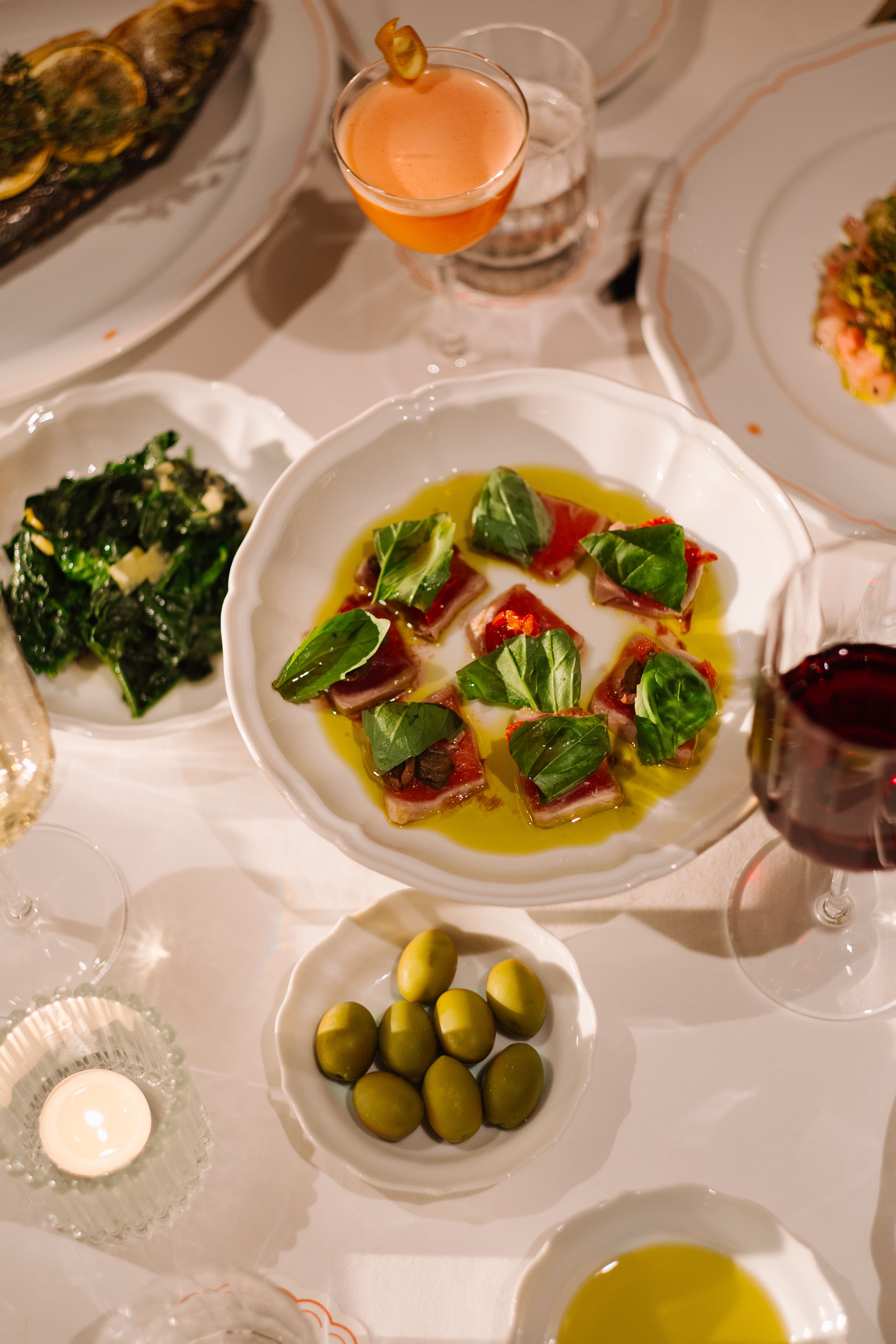 Overhead view of an elegant dining table set with white scalloped plates, featuring tuna carpaccio topped with basil and olive oil at center, a small bowl of green olives, sautéed greens, a citrus-garnished cocktail, red wine, and a softly lit candle creating a warm, refined atmosphere.