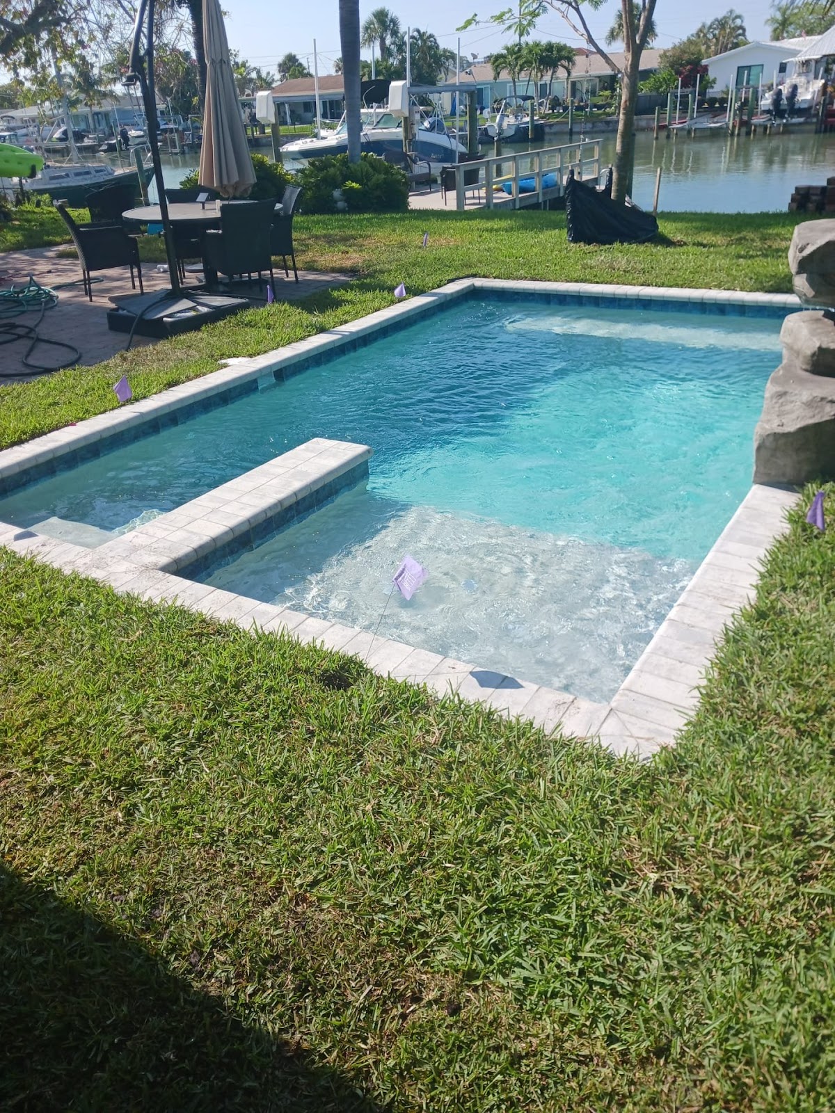 Rectangular backyard swimming pool with an attached spa, light stone deck, manicured landscaping, and waterfront setting.