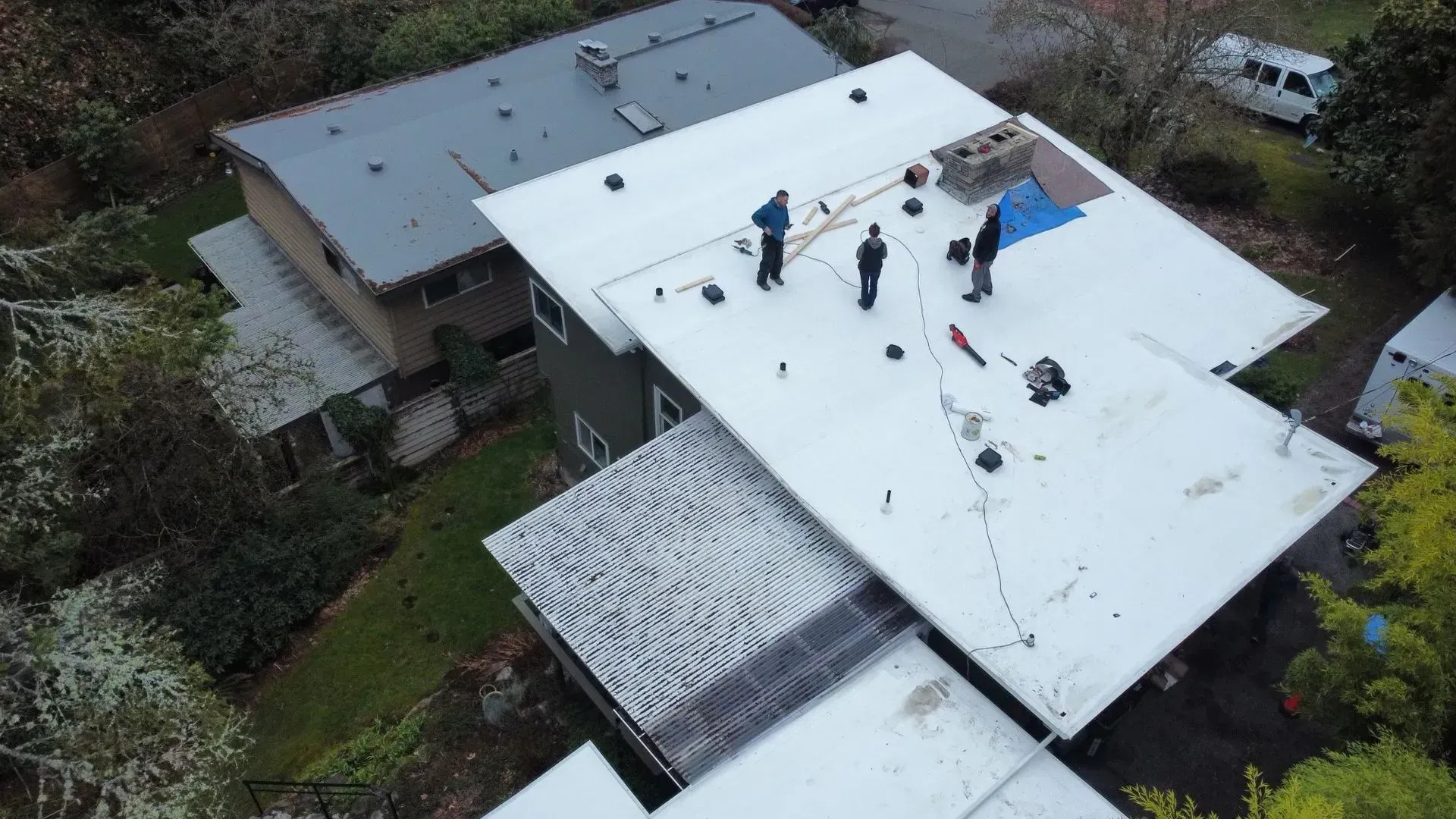 An aerial view of a two-story house with a large, white flat roof. Three workers are standing on the roof with tools and materials, working on what appears to be a new installation.
