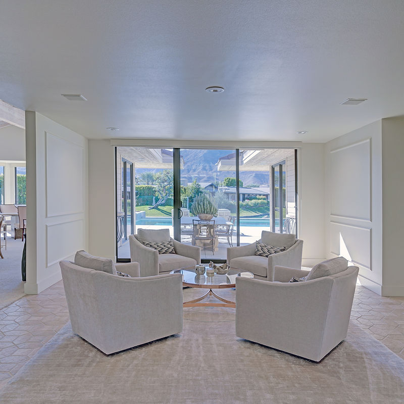A living room interior with four plush, light-colored armchairs arranged in a circle around a glass-top coffee table. Large glass doors in the background open to a patio with a swimming pool and mountains in the distance. The room features minimalist decor, with light walls and a neutral-toned rug.