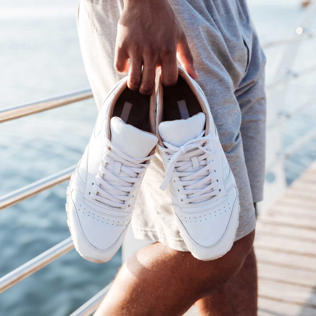 A person holding a pair of white leather sneakers with white laces in front of a blurred background of water and a wooden deck. The person is wearing grey athletic shorts. The sneakers appear to be clean and in good condition.