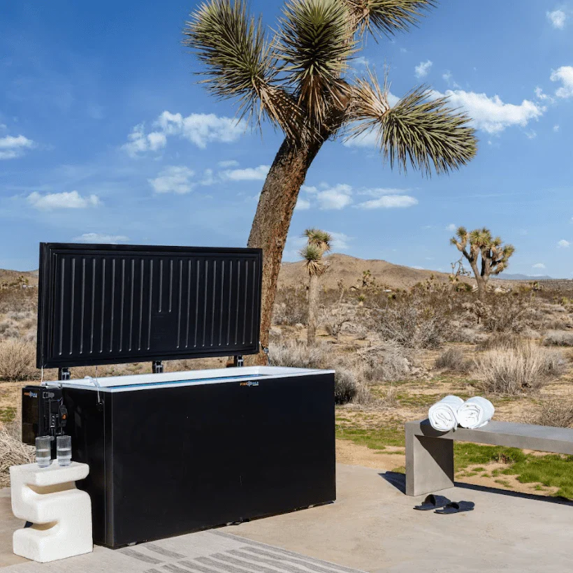 A black "All-In-One" ice bath sits outdoors in a desert landscape, with a large Joshua tree to its left and more desert vegetation in the background under a bright blue sky with scattered clouds. The lid of the ice bath is open, revealing the white interior. To the left of the ice bath is a modern white side table with two glasses of water, and to the right, a concrete bench with two rolled white towels and a pair of dark flip-flops.