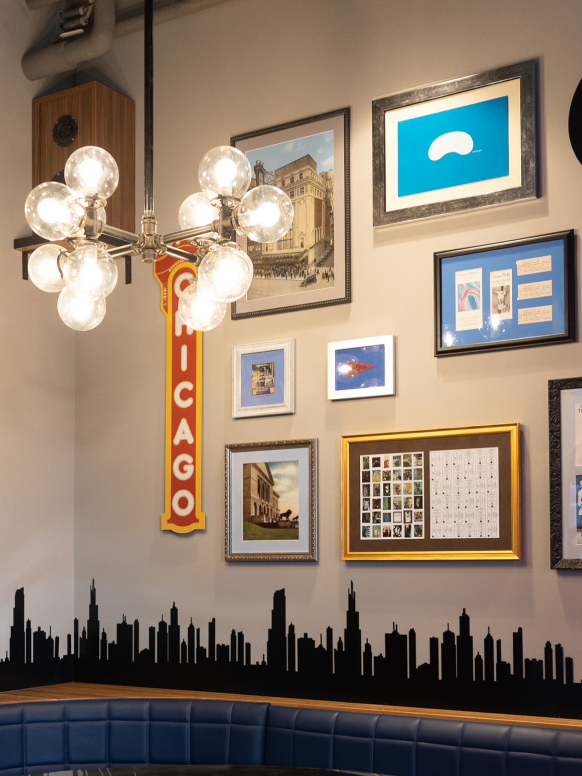 A corner of an interior space featuring a gallery wall of Chicago-themed decor. A modern, industrial-style chandelier with multiple spherical bulbs hangs to the left of a vertical, vintage-style marquee sign reading "CHICAGO" in red letters on a yellow background. The surrounding framed artwork includes a graphic of the Cloud Gate "Bean" sculpture, vintage street scenes, and a photo of the Art Institute lions. A black silhouette decal of the Chicago skyline runs along the bottom of the wall, positioned just above the back of a navy blue, tufted leather booth.