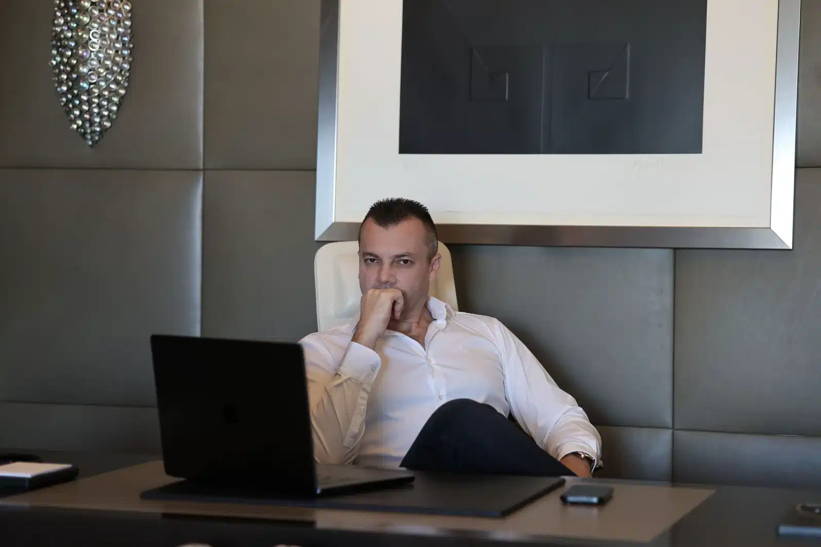 A man in a white dress shirt sits at a desk with a laptop in front of him. He is resting his chin on his hand and looking at the camera. The wall behind him is paneled in a diamond pattern and a large framed picture hangs on the wall. A decorative light fixture hangs on the left side of the image.