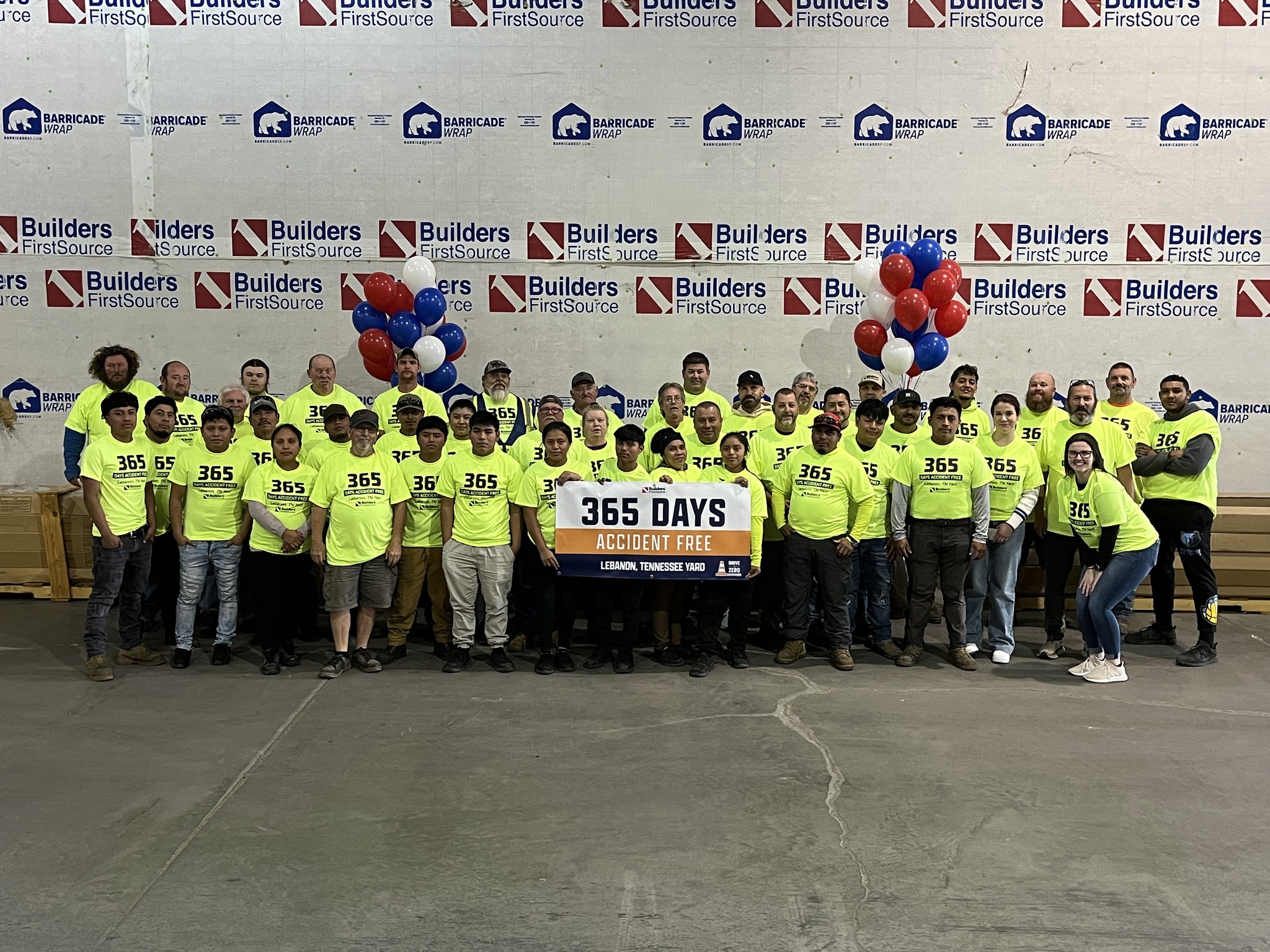 A group photo of Builders FirstSource employees at the Lebanon, Tennessee yard celebrating a milestone of 365 days accident-free. The team is wearing bright yellow shirts with "365" printed on them, standing in front of a backdrop featuring the Builders FirstSource and Barricade Wrap logos. Red, white, and blue balloons add a festive touch to the scene, with a banner prominently displaying the achievement.