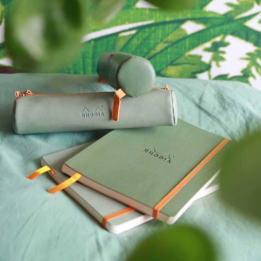 A professionally styled product image by Tabula Rasa Social Stationers, featuring a collection of Rhodia stationery. The display showcases two sage green notebooks with signature orange elastic bands and ribbon bookmarks, alongside two coordinating sage green pencil cases with orange zipper pulls. This visual highlights Rhodia's commitment to French quality and minimalist design, emphasizing products crafted for a smooth writing experience, ideal for professionals and creatives seeking durable and sophisticated tools.